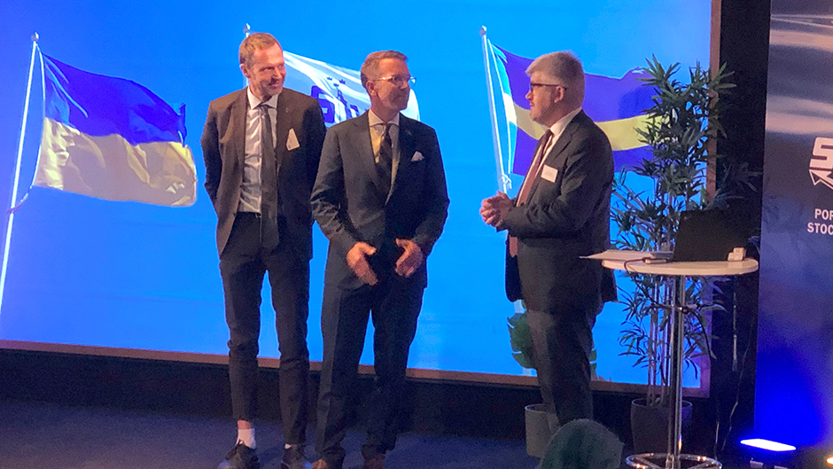 Tre män på en scen. Ukarinska, svenska samt Stockholms Hamnars flagga i bakgrunden.